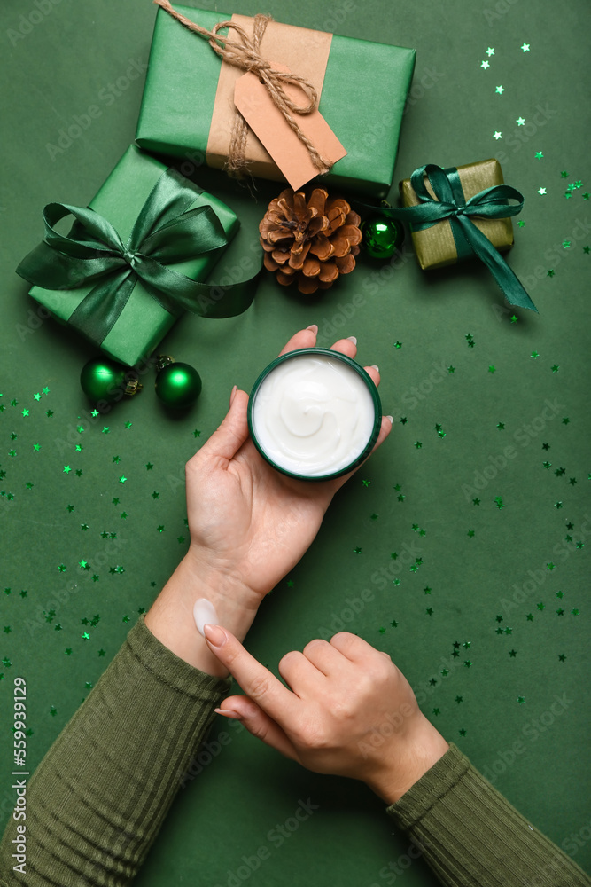 Female hands with jar of cream, Christmas gifts and decorations on green background