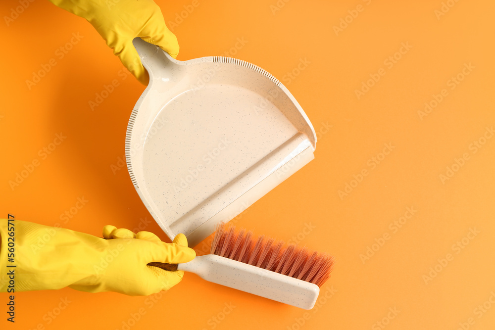 Female hands in rubber gloves with dustpan and brush on orange background