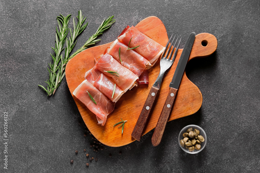 Wooden board with rolled slices of tasty ham, capers and rosemary on dark background