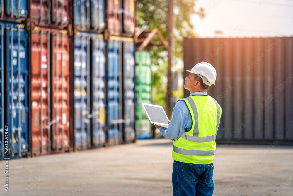 Engineer or foreman holding laptop and wears PPE checking container storage with cargo container bac