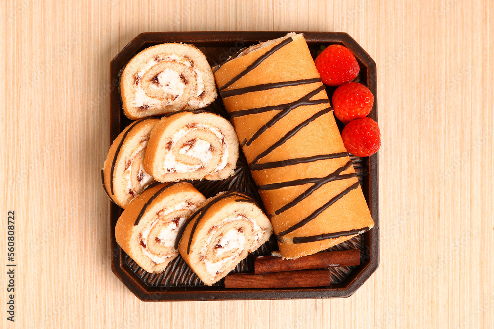 Board with pieces of tasty sponge cake roll, cinnamon and strawberry on wooden background