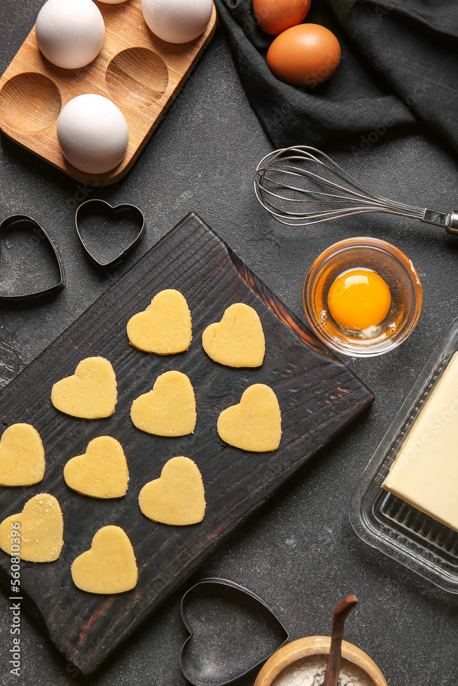 Wooden board with raw heart shaped cookies, ingredients and cutters on dark background. Valentines D