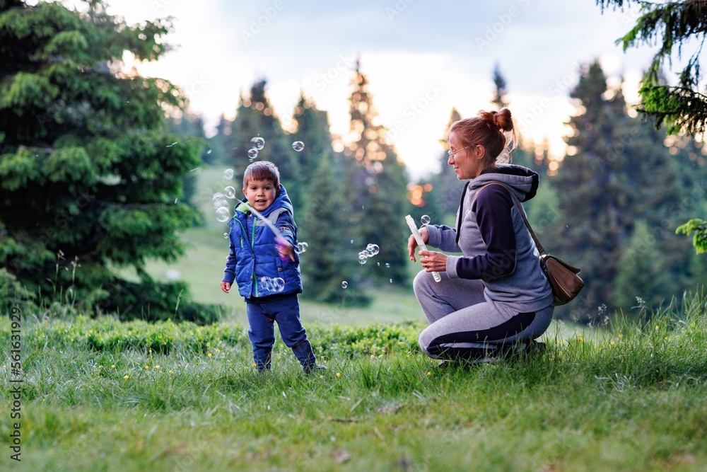 Mother plays with son on a lawn and teaches him to blow soap bubbles against the backdrop of forest