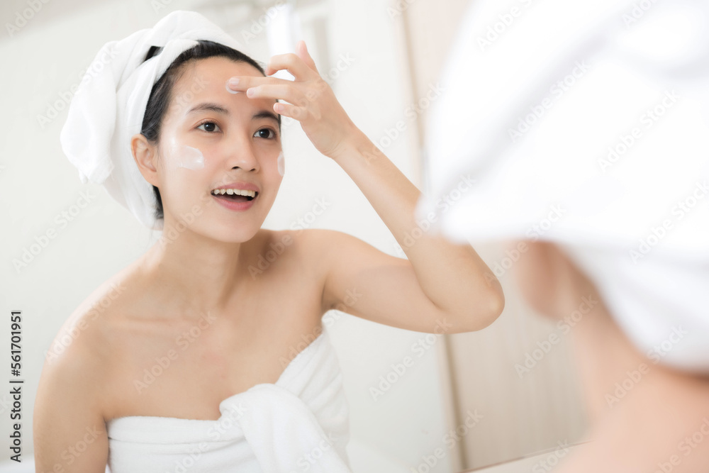 Happy young Asian woman applying face lotions while wearing a towel and touching her face in bathroo