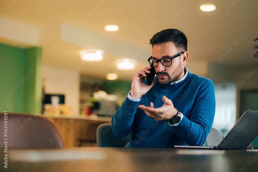 Male employer sitting in front of the laptop, using a mobile phone, with glasses on.