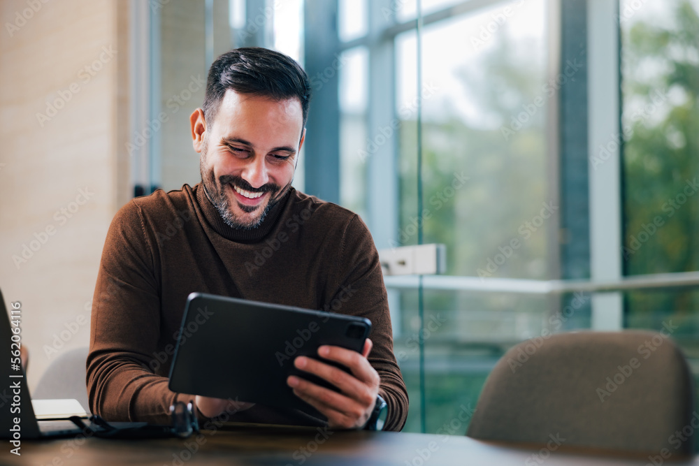 Smiling male employer using a digital tablet, taking a work break, sitting at the office.