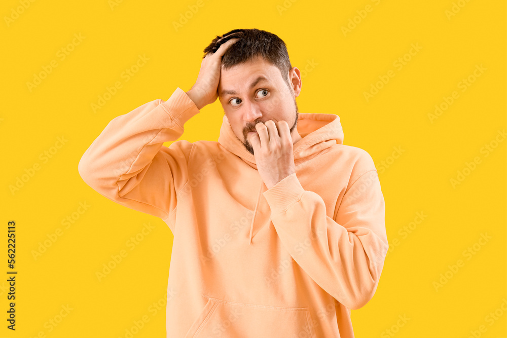 Handsome man in hoodie biting nails on yellow background