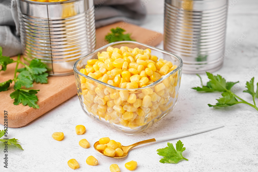 Bowl with corn kernels, tin cans, spoon and parsley on white table