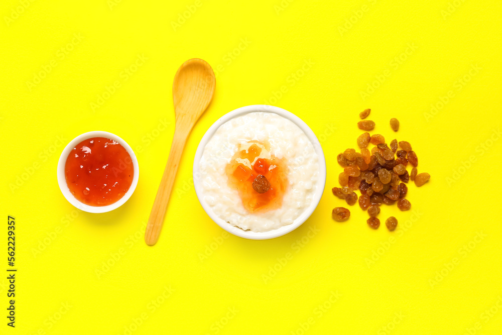 Bowl with delicious rice pudding, jam, raisins and spoon on yellow background