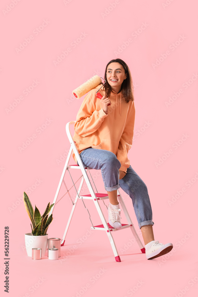 Young woman with paint roller, ladder, cans and houseplant on pink background