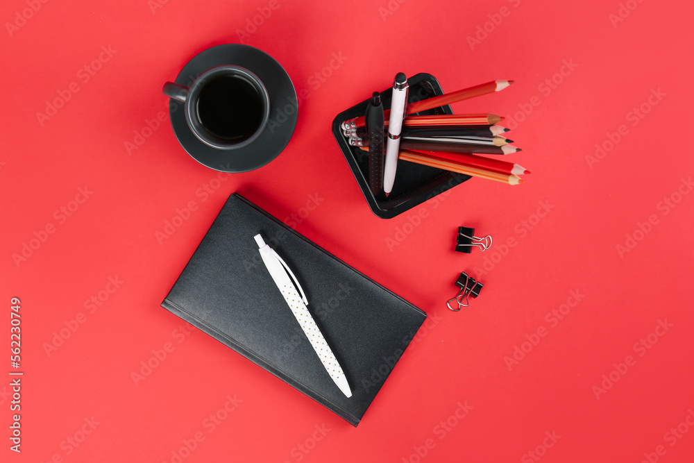 Notebook, stationery holder,  paperclips and cup on red background