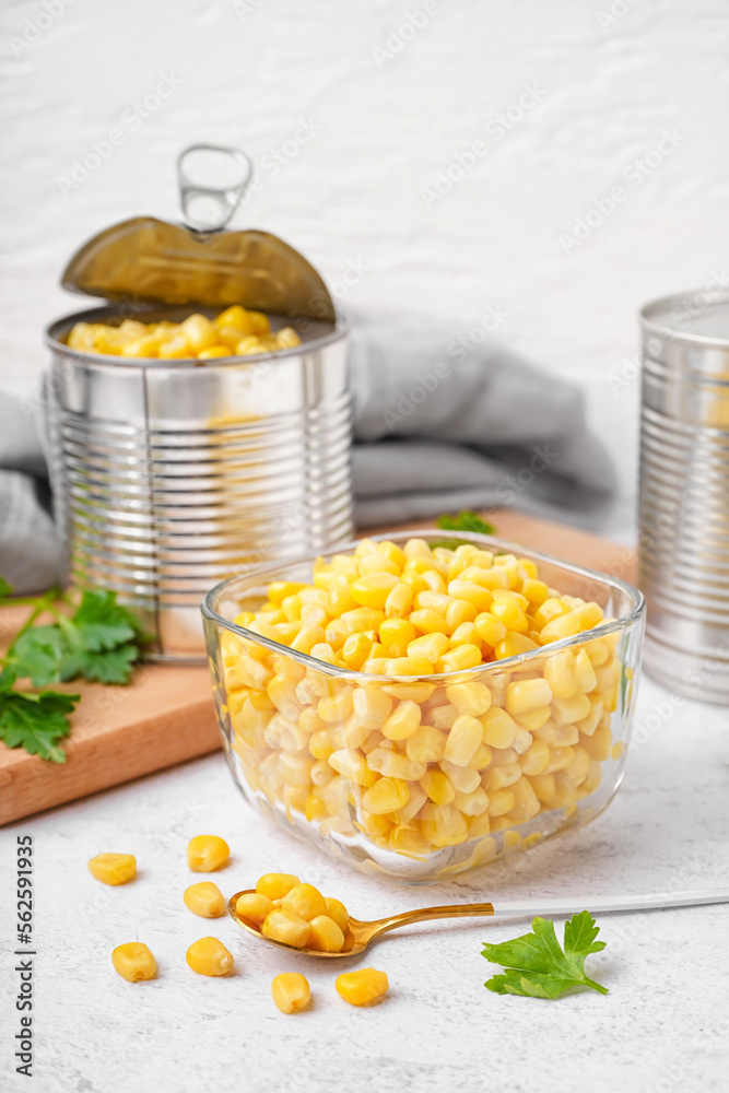 Bowl with corn kernels, tin cans, spoon and parsley on white table