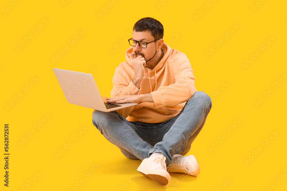 Handsome man with laptop biting nails on yellow background