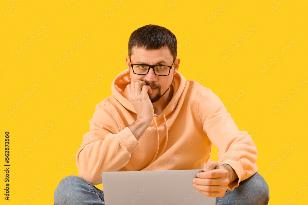 Handsome man with laptop biting nails on yellow background