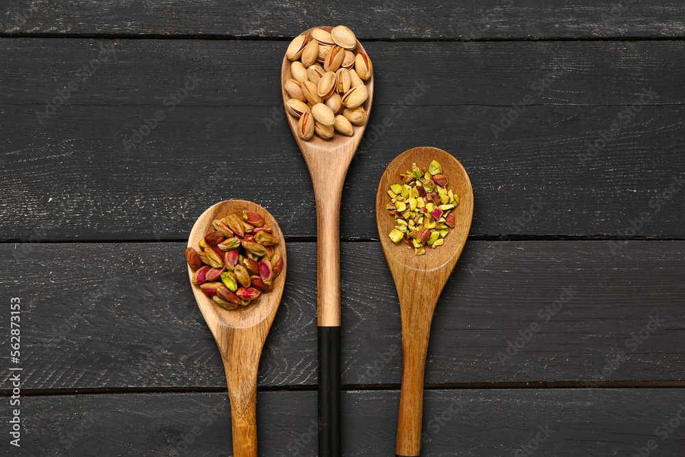 Spoons with unpeeled, peeled and sliced pistachios on black wooden background