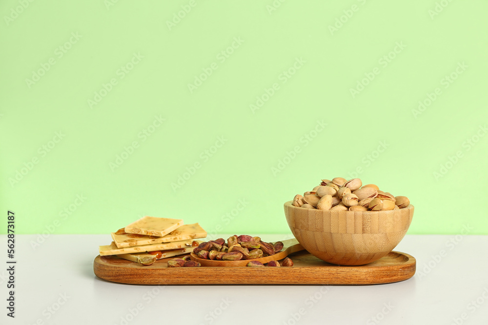 Board with bowl of tasty pistachio nuts and chocolate on green background