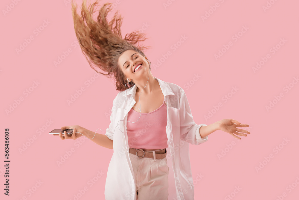 Young woman in earphones with mobile phone dancing on pink background