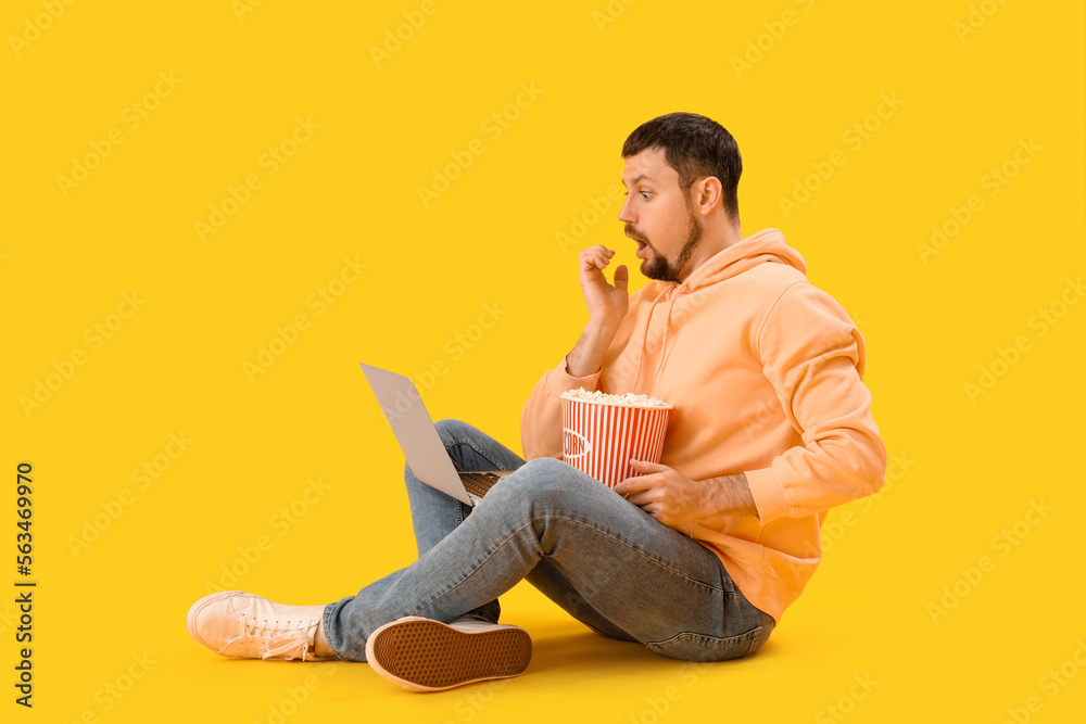 Handsome man with laptop biting nails on yellow background