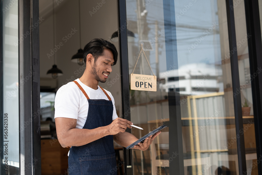 Portrait of smiling owner standing at his restaurant gate with open signboard. Young entrepreneur le