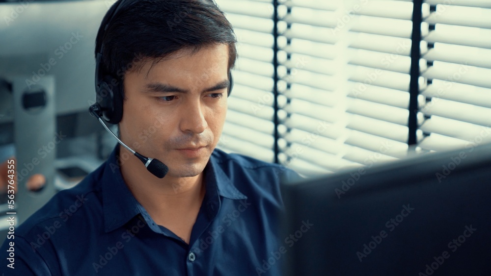 Young competent asian male call center agent working at his computer while simultaneously speaking w