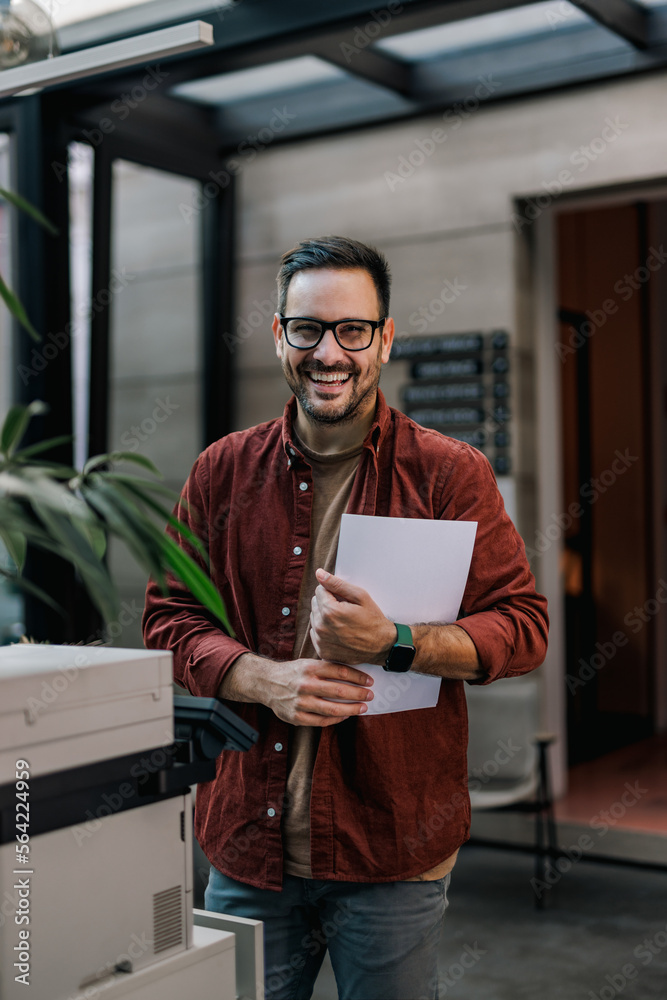 Smiling businessman holding some documents, standing at the office, in front of a scanner.