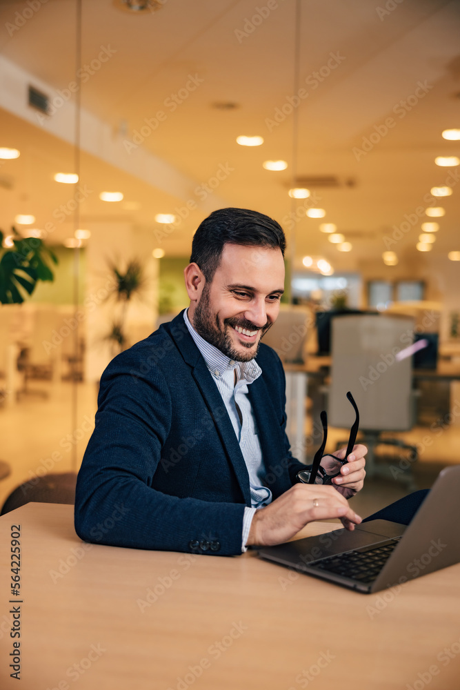 Smiling man working over the laptop, sitting at the office, holding glasses, elegantly dressed.