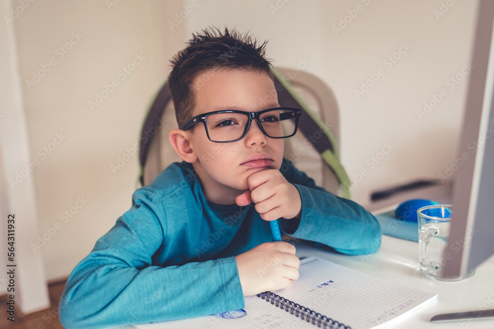 Serious dreamy little boy with glasses sitting at desk in his room, while doing homework.