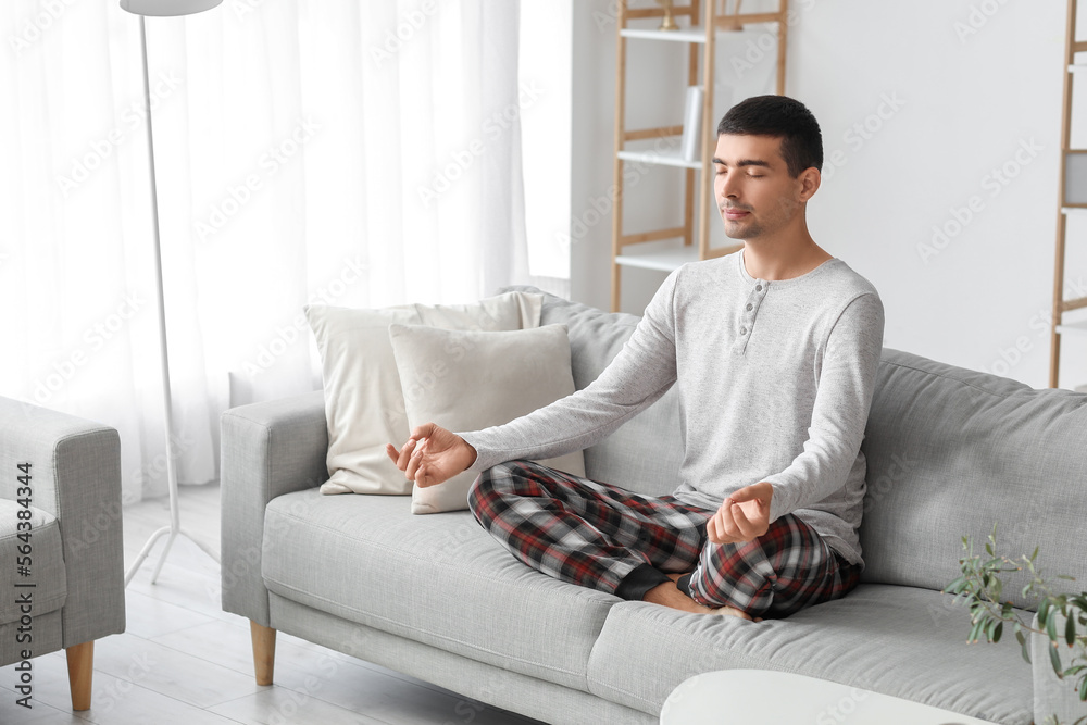 Young man in pajamas meditating on sofa at home