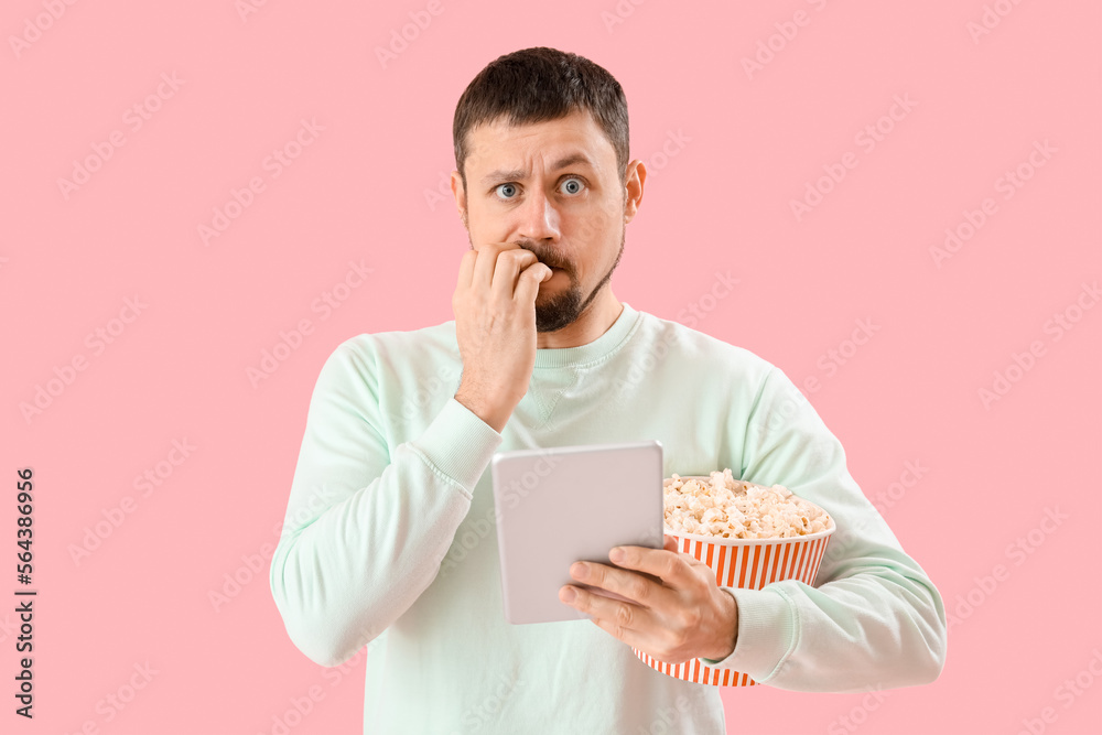 Handsome man with tablet computer and popcorn biting nails on pink background