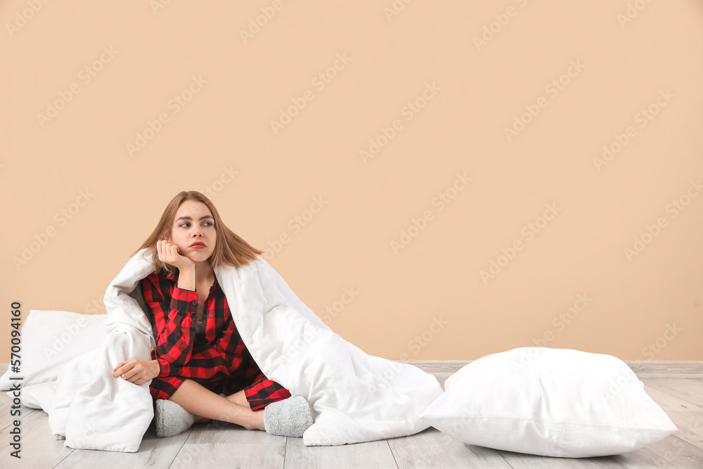 Upset young woman in pajamas with blanket sitting near beige wall