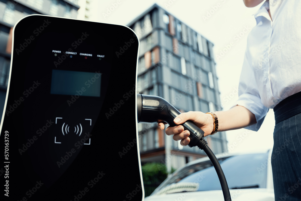 Focus EV charger plug and electric car at public charging station with blur progressive businesswoma