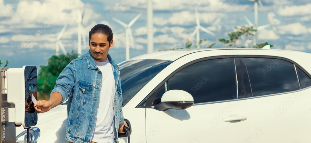 Progressive man with his electric car, EV car recharging energy from charging station on green field