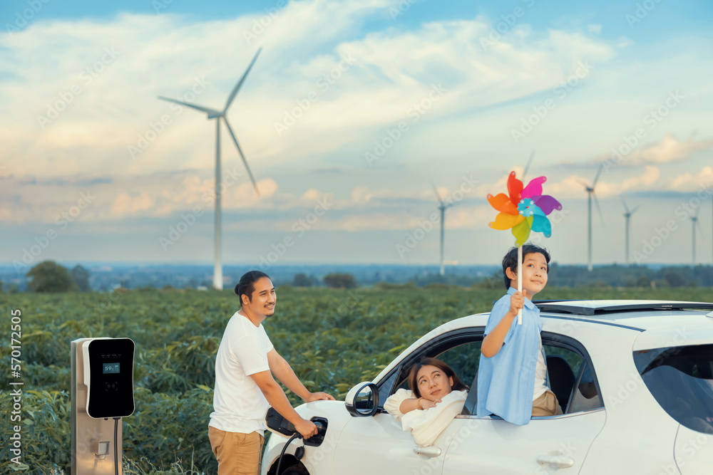 Concept of progressive happy family enjoying their time at wind farm with electric vehicle. Electric