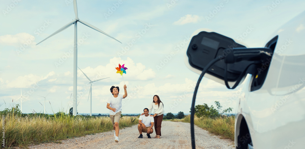 Concept of progressive happy family holding windmill toy and relax at wind farm with electric vehicl