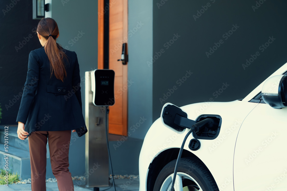 Progressive woman install cable plug to her electric car with home charging station. Concept of the 