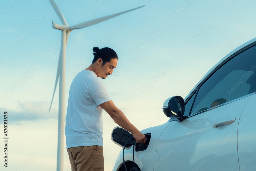 Progressive man with his electric car, EV car recharging energy from charging station on green field