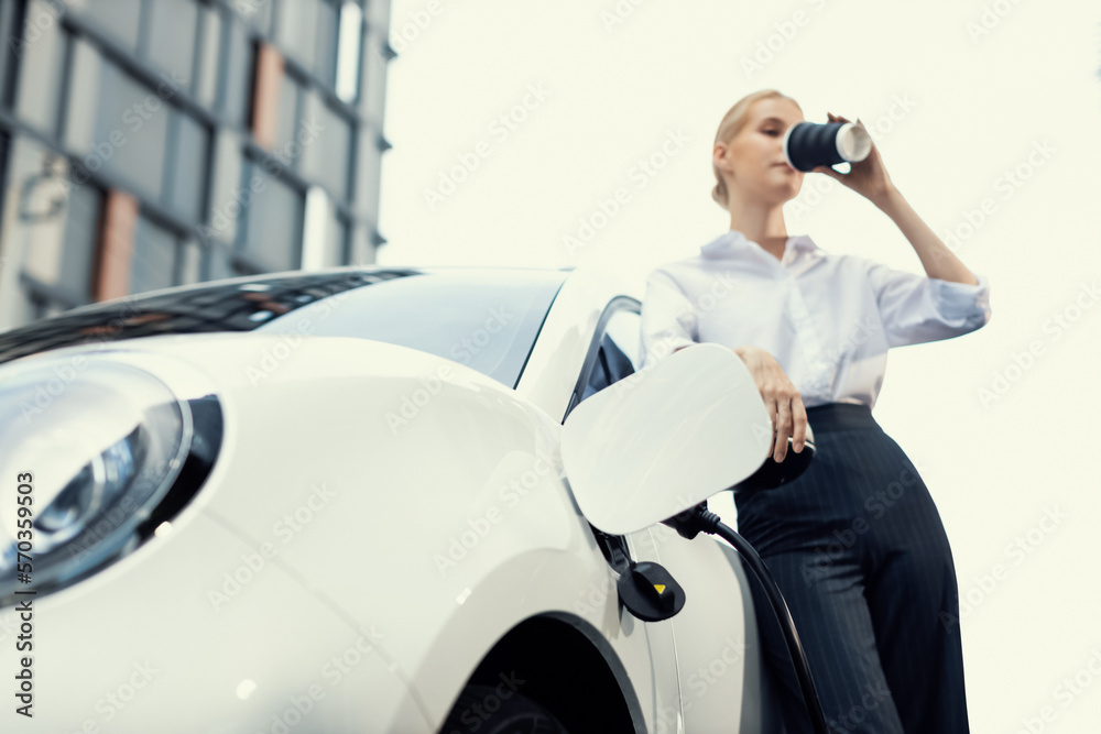 Progressive businesswoman with electric car and charging station before driving around city center. 