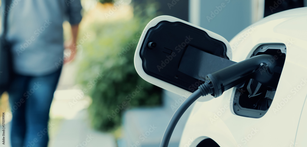 Focus electric car charging at home charging station with blurred progressive man walking in the bac