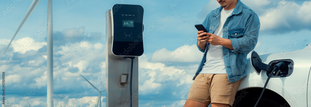 Progressive man with his electric car, EV car recharging energy from charging station on green field