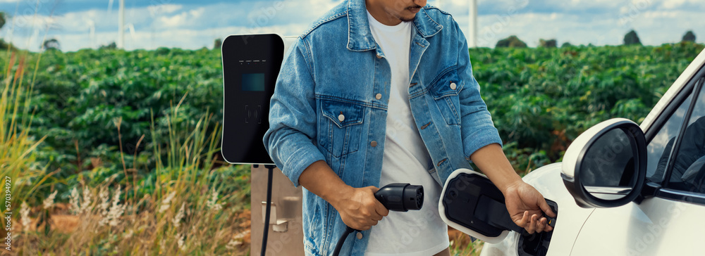 Progressive man with his electric car, EV car recharging energy from charging station on green field