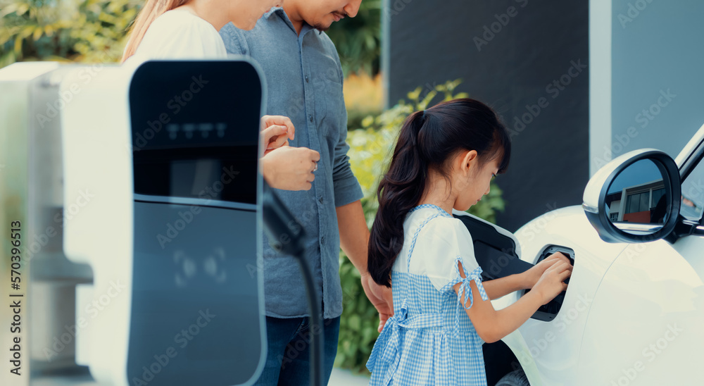 Progressive young parents and daughter with electric vehicle and home charging station. Green and cl