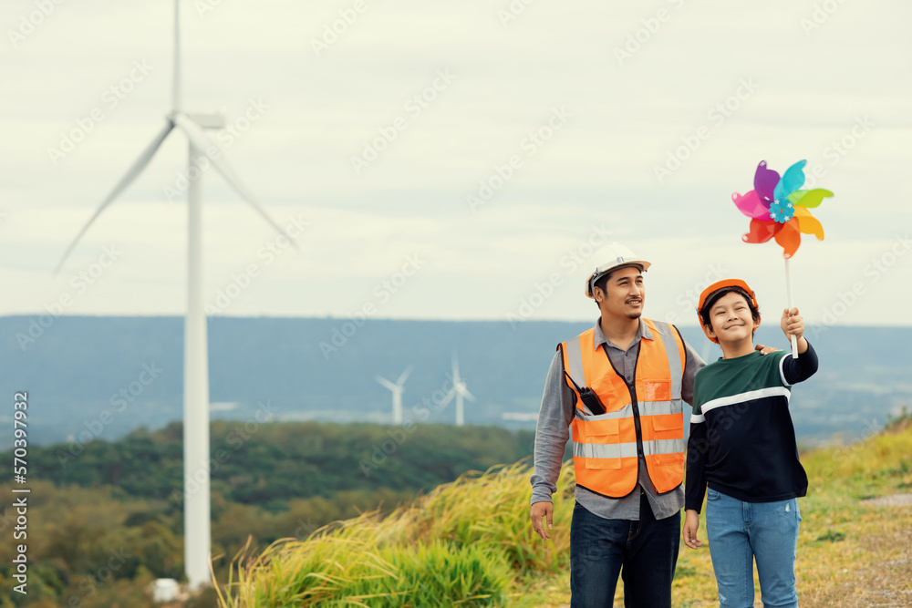 Engineer with his son holding windmill toy on a wind farm atop a hill or mountain. Progressive ideal