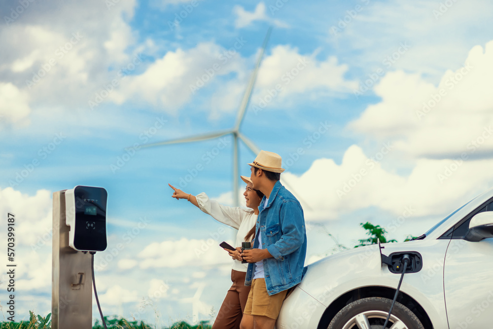 Concept of progressive happy young couple enjoying their time at windmill farm with electric vehicle