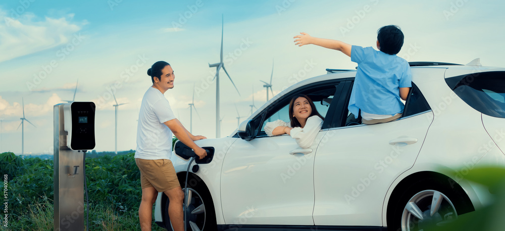 Concept of progressive happy family enjoying their time at wind farm with electric vehicle. Electric