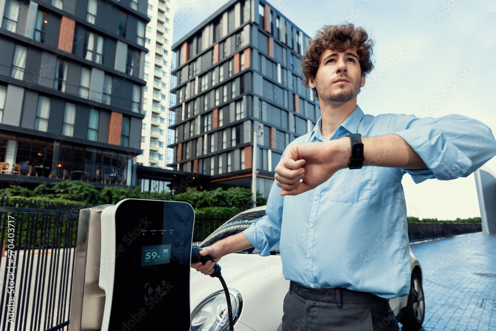 Businessman with smartwatch at modern charging station for electric vehicle with background of resid