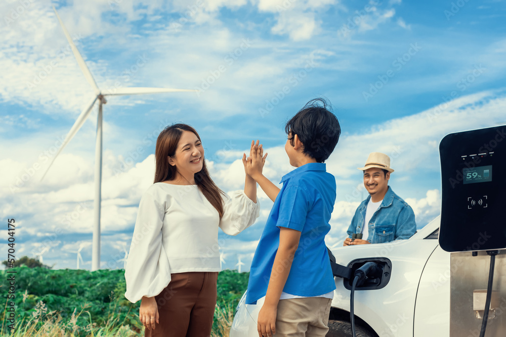 Concept of progressive happy family enjoying their time at wind farm with electric vehicle. Electric