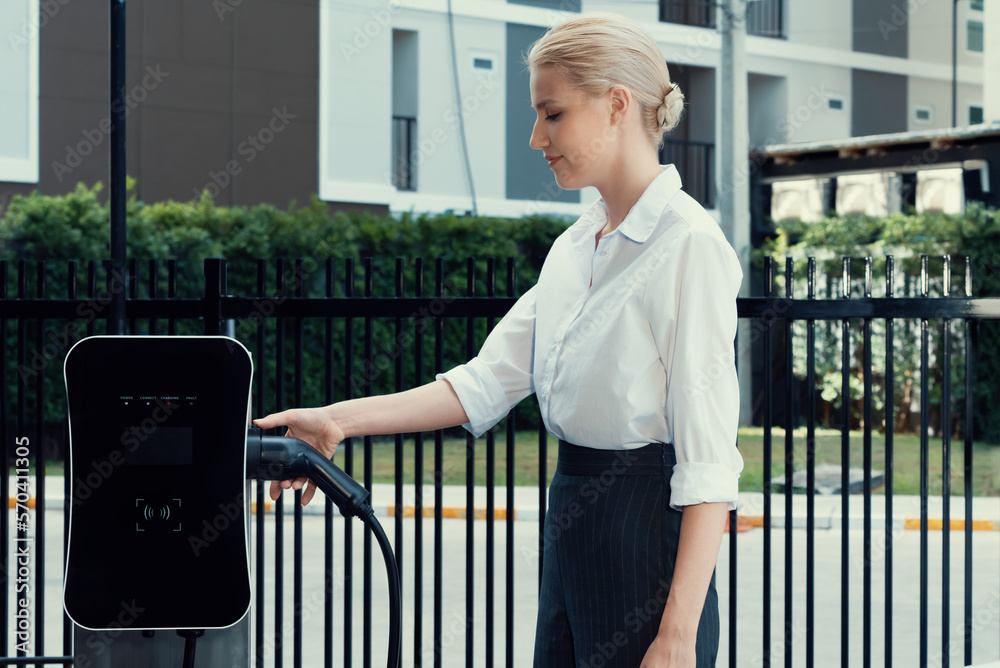 Progressive businesswoman insert charger plug from charging station to her electric vehicle with apa