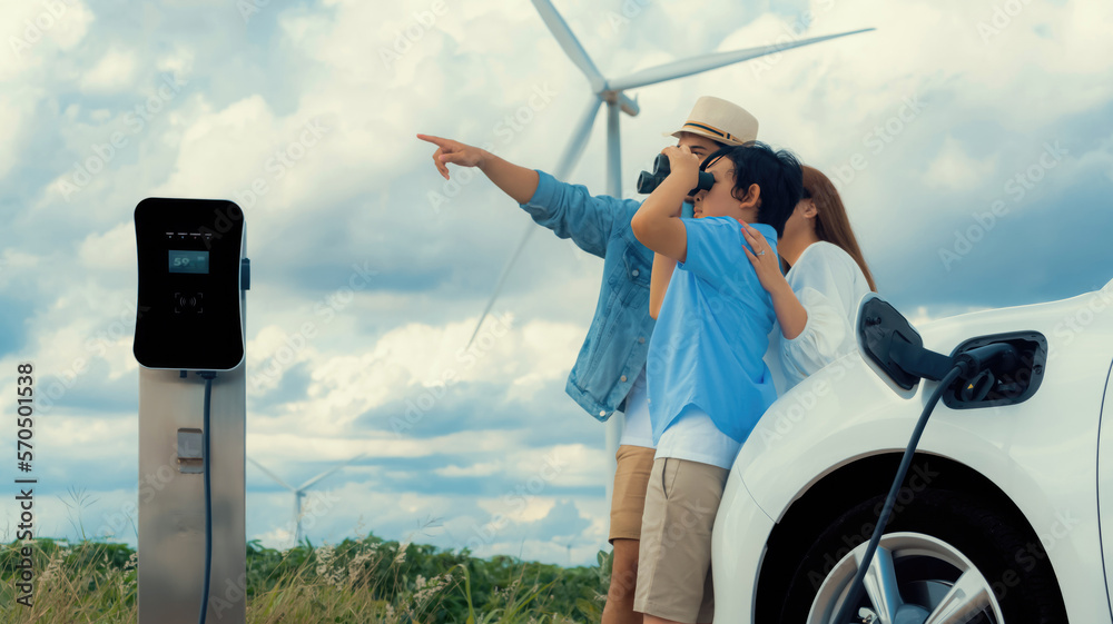 Concept of progressive happy family enjoying their time at wind farm with electric vehicle. Electric