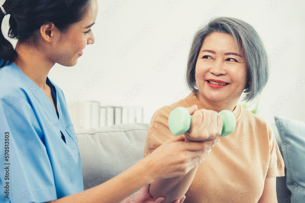 Caregiver helping contented senior woman exercise with dumbbell at home. Rehab for elder, senior med