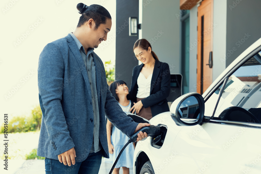 Progressive young parents and daughter with electric vehicle and home charging station. Green and cl